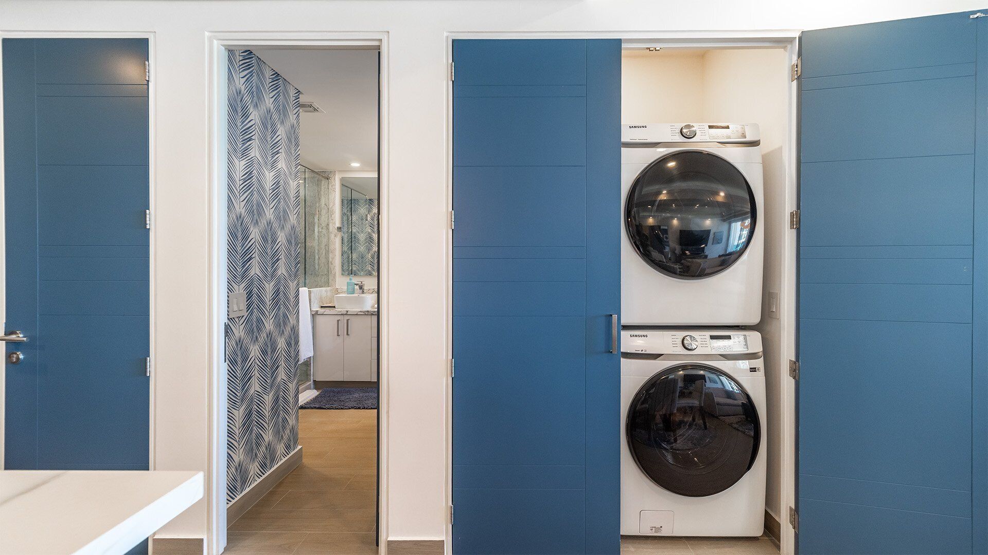 Une buanderie équipée de deux machines à laver Samsung superposées, une salle de bain avec un miroir et un lavabo, et un mur décoré d’un motif.