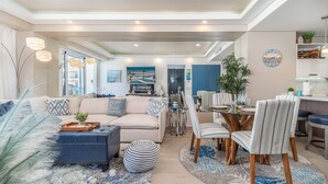 A modern living room with a sofa, ottoman, and striped chairs. There is a dining area with a table and chairs. A large painting is on the wall, and a plant is in the corner. - Sublime 1 Bedroom Condo Encantame Towers Verano 403 (Puerto Peñasco)