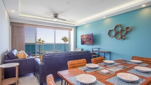 Ein modernes Wohnzimmer mit großem Fenster zum Meer, einer Essecke mit Holztisch für vier Personen und einer blauen Akzentwand mit sechsecklicher Dekoration.