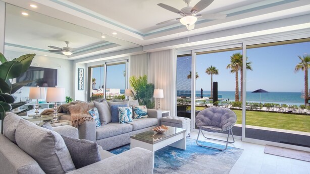 A modern living room with a grey sofa, coffee table, and armchair, large windows with a view of the ocean and palm trees, and ceiling fans. - Lovely 2 Bedroom Condo Encantame Towers Velero 104 (Puerto Peñasco)