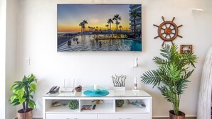 A living room with a large painting of a beach scene, a wooden ship wheel decoration, and a white entertainment unit with various items. - Lovely 2 Bedroom Condo Encantame Towers Velero 104 (Puerto Peñasco)