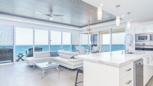 A modern interior with a large kitchen, dining area, and seating by a large window overlooking the sea.