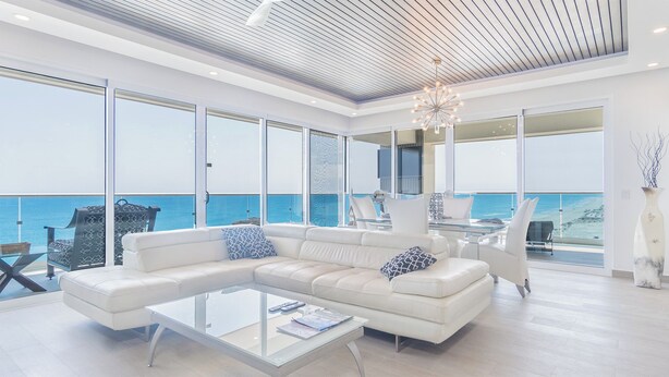 A modern living room with a large white sectional sofa, a glass coffee table, and a view of the ocean through large windows.
