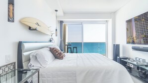A hotel room with a bed, a surfboard on the wall, a balcony with a view of the sea, and a desk with a computer.
