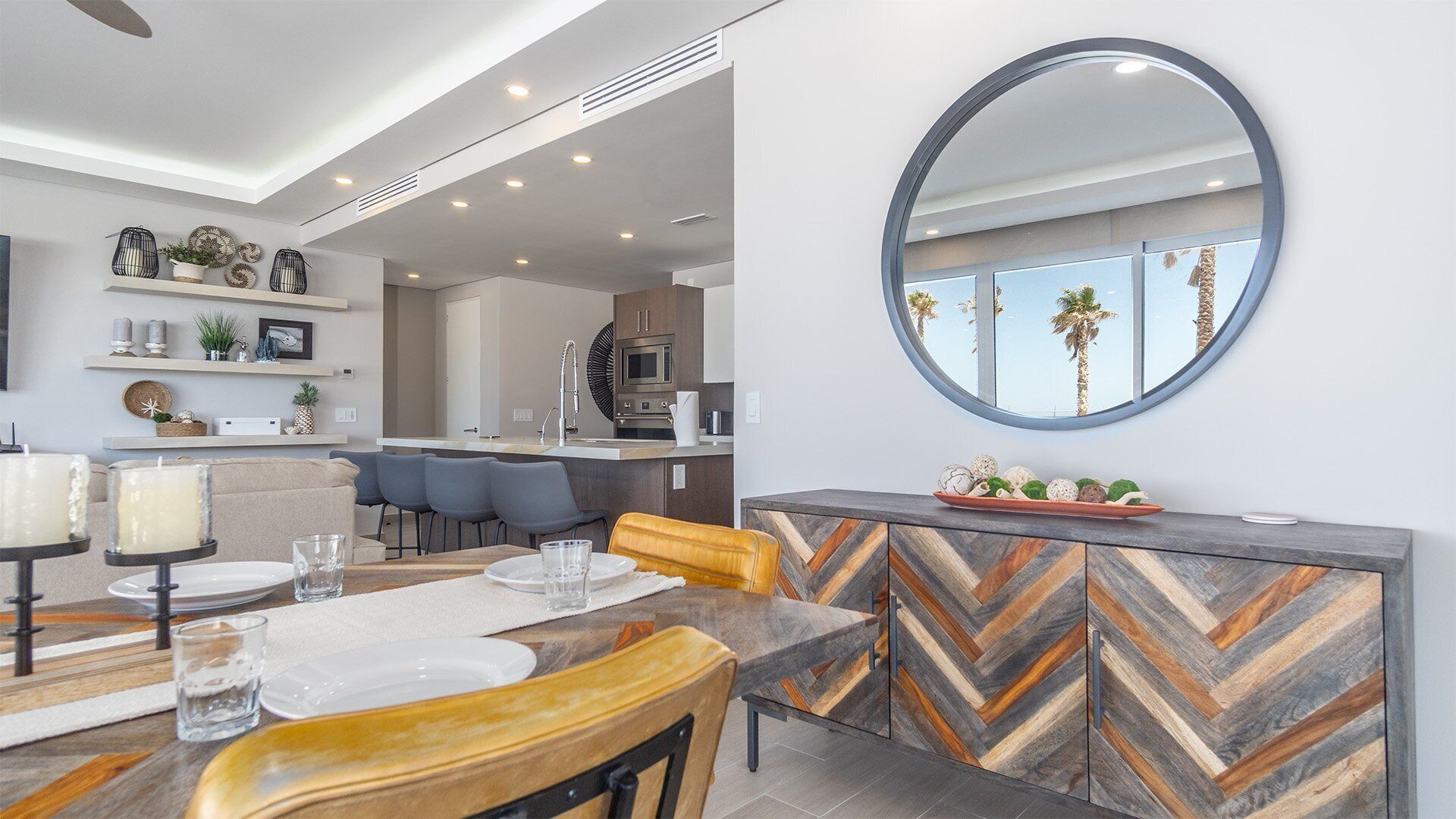 A modern dining area with a herringbone-patterned cabinet, a round mirror, and a view of the kitchen in the background.