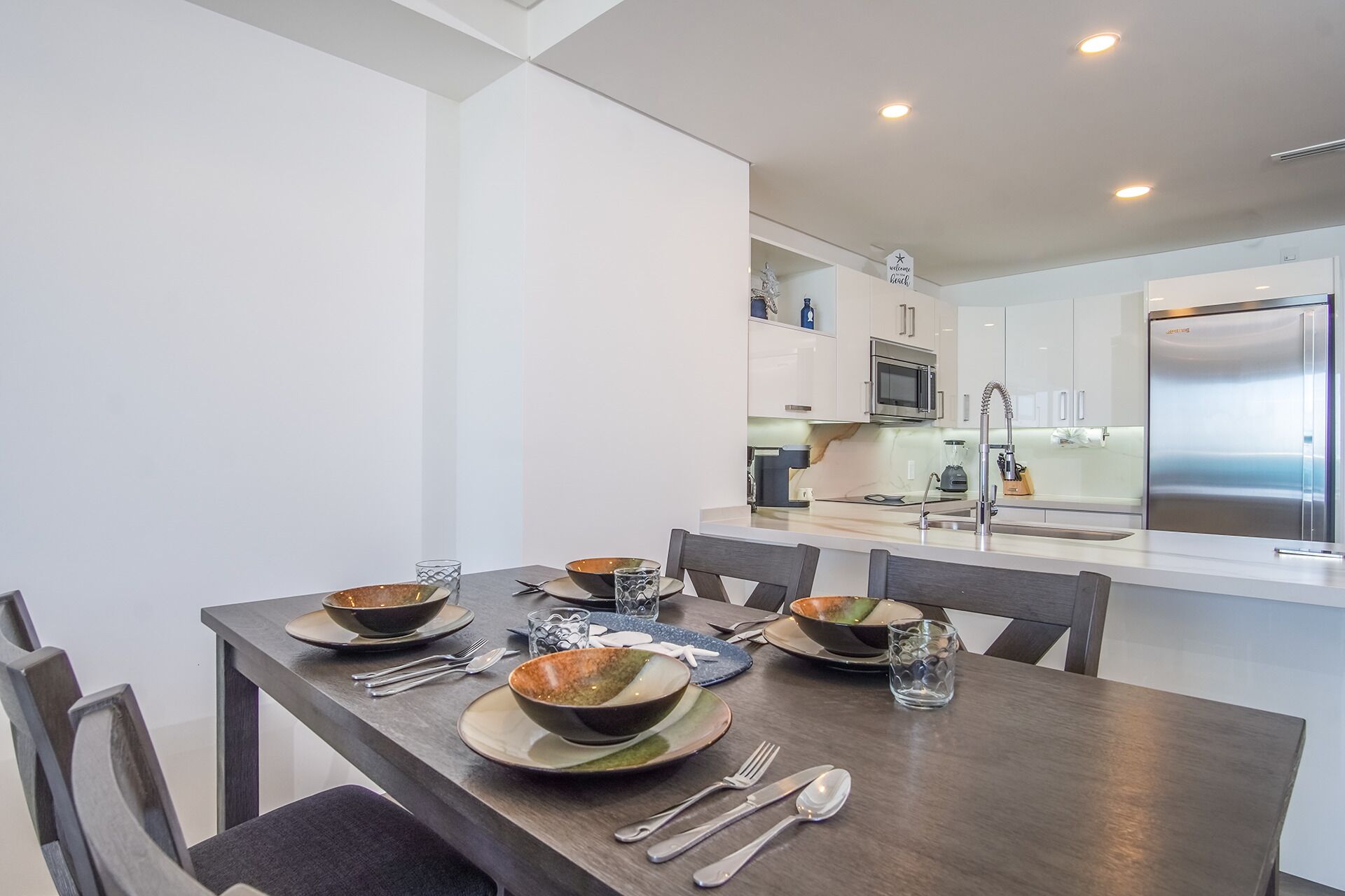 Un comedor moderno con una mesa de madera dispuesta para cuatro personas, una cocina al fondo con armarios blancos y electrodomésticos de acero inoxidable.
