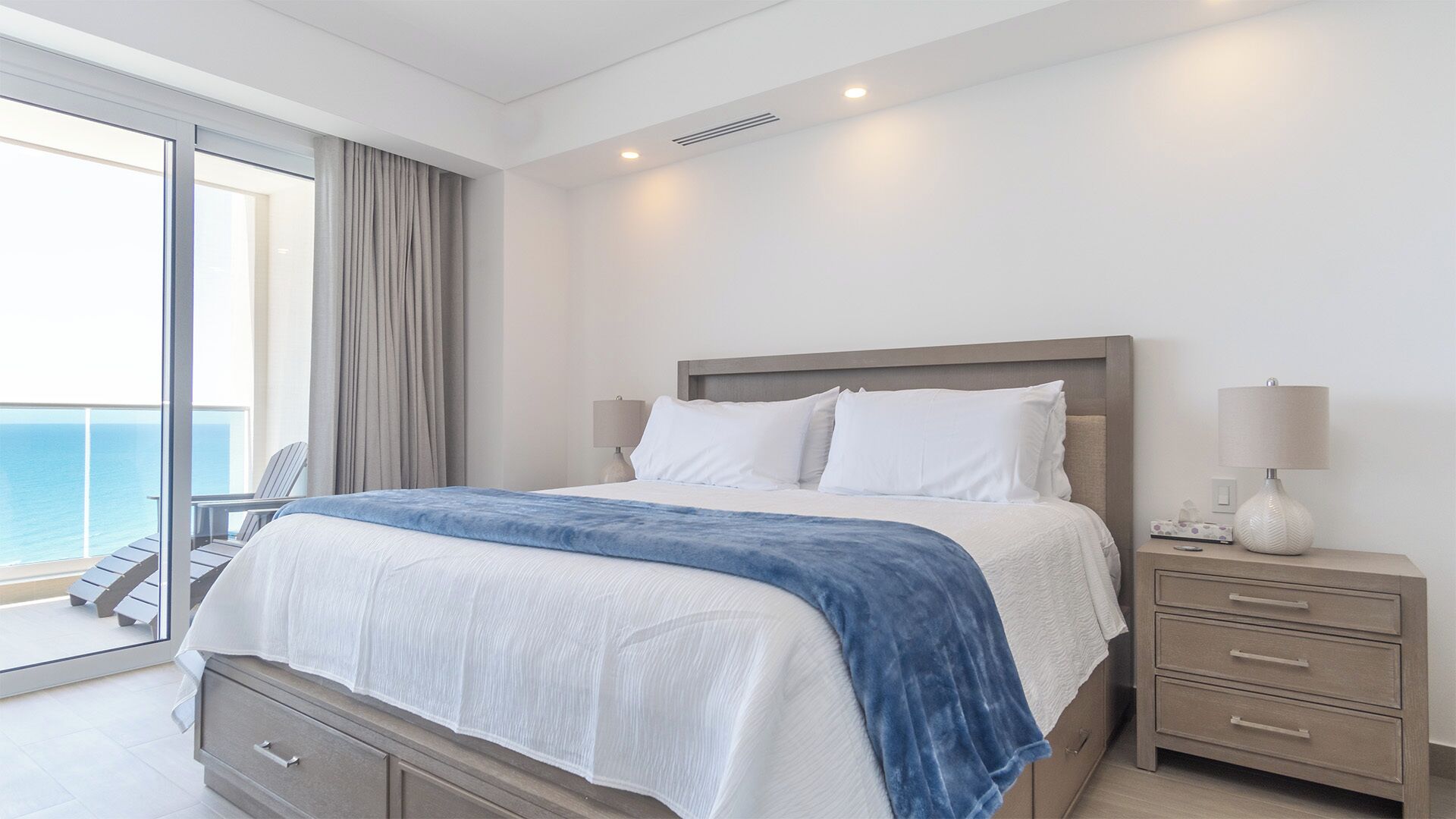 Una habitación de hotel moderna con una cama grande, una mesita de noche con lámpara, un aparador y vistas al océano a través de una puerta corrediza de vidrio.