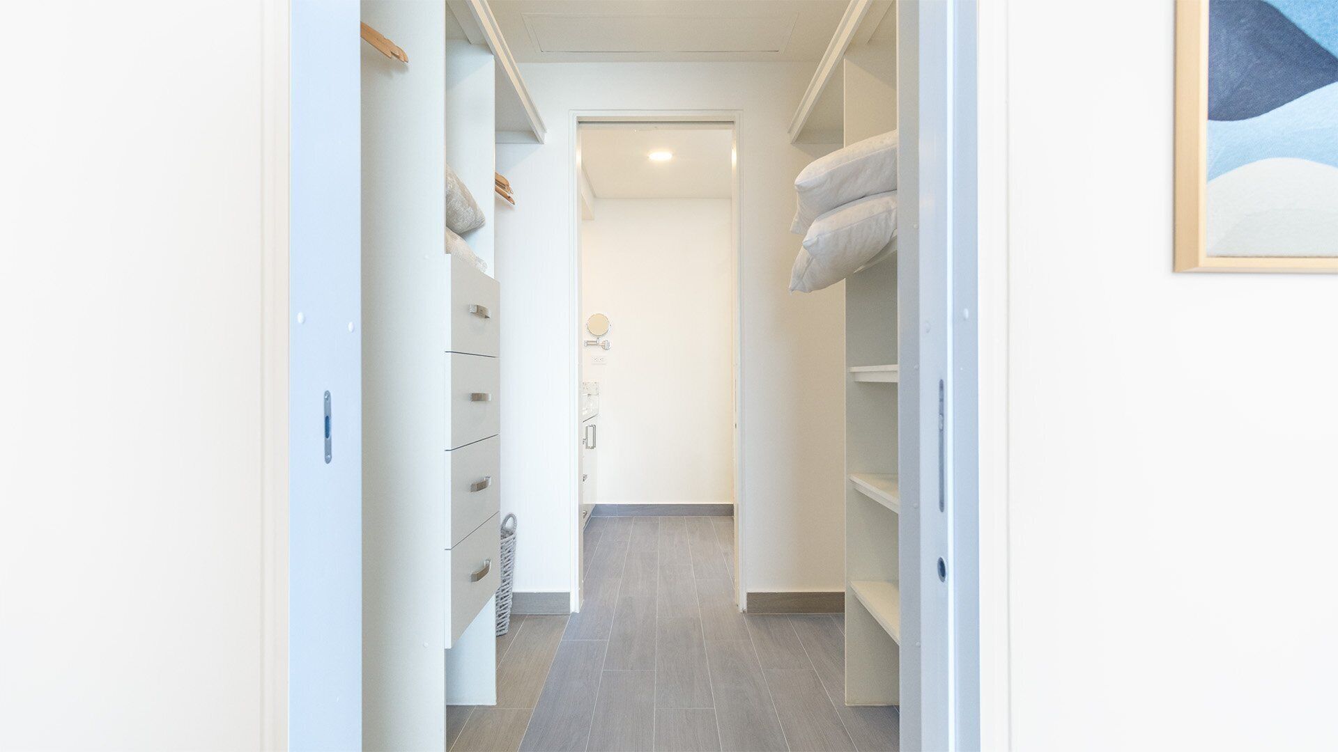 A narrow hallway with white walls and grey flooring, leading to a room with a door.