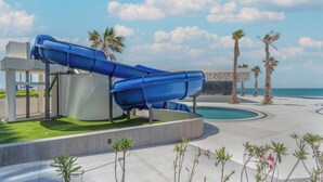 Ein Wasserpark mit zwei blauen Rutschen, einem Pool, Palmen und blauem Himmel.