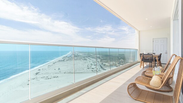 Ein Balkon mit Glastrennwand blickt auf einen Strand. Korbstühle und ein Tisch mit einem Glas Orangensaft stehen bereit.