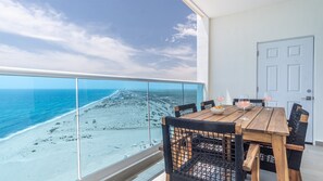 Een balkon met een houten tafel en stoelen, een glazen hekwerk, en uitzicht op een strand en zee.