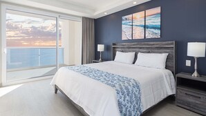 Ein Hotelzimmer mit einem großen Bett, zwei Nachttischen mit Lampen, Meerblick durch einen Balkon und einem Sonnenuntergangsbild an der Wand.