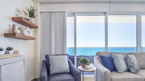 Un salon moderne avec un canapé gris, un fauteuil bleu et une petite table ronde agrémentée d’une plante. On y trouve des étagères en bois ornées d’objets décoratifs et une grande fenêtre donnant sur la mer.