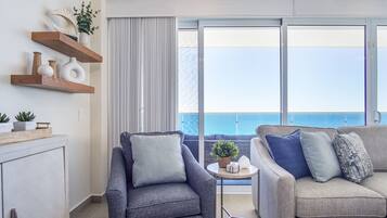 A modern living room with a grey sofa, a blue armchair, and a small round table with a plant. There are wooden shelves with decorative items and a large window with a view of the sea.