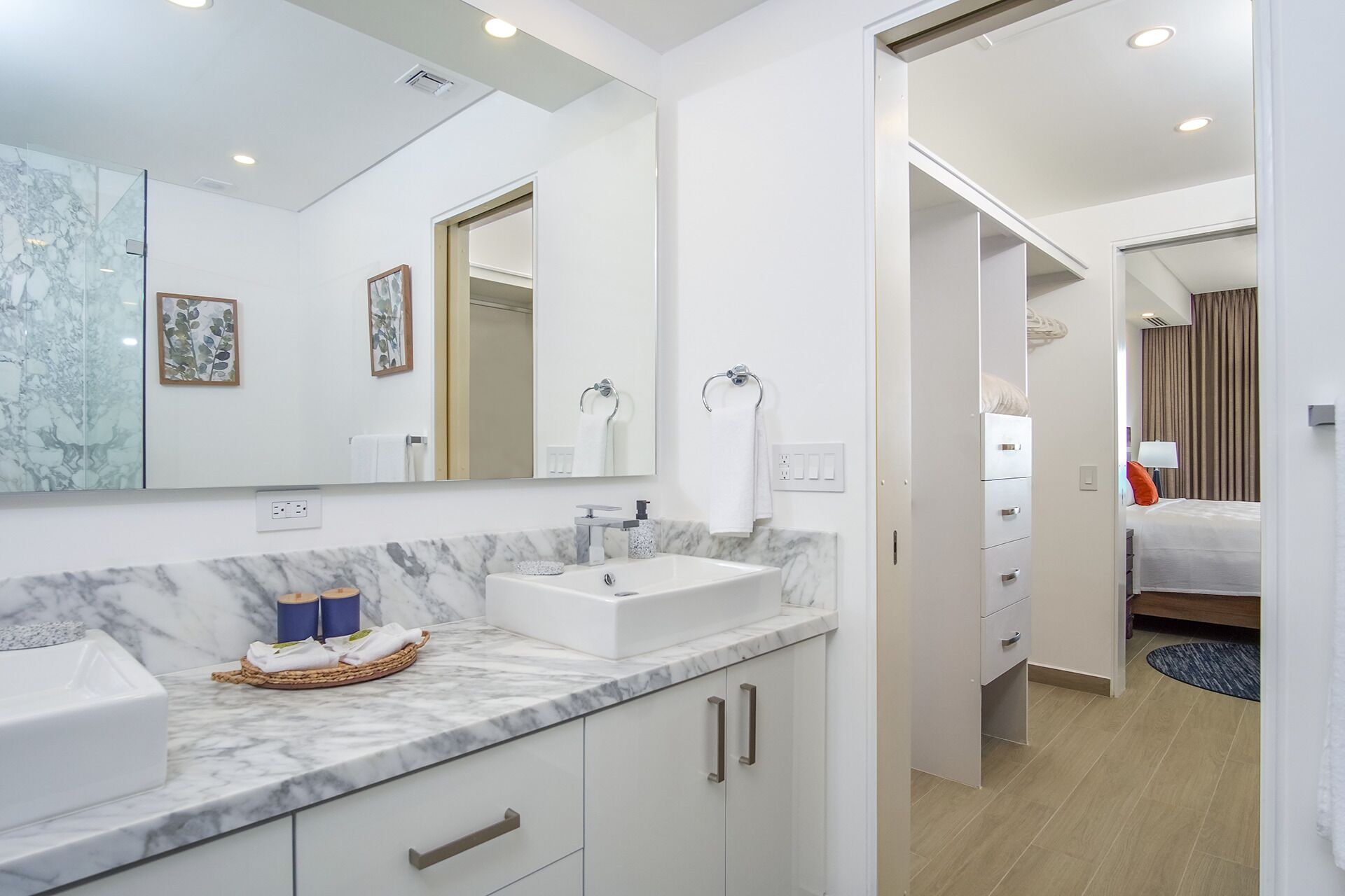 A modern bathroom with a marble countertop, a sink, and a large mirror. There is a door leading to a bedroom with a bed and a nightstand.