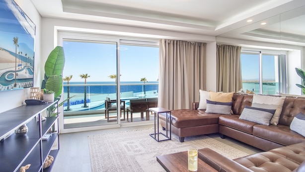 Ein geräumiges Wohnzimmer mit einem großen Fenster und Blick auf den Ozean und Palmen. Es gibt ein braunes Ledersofa mit Kissen und einen kleinen Holztisch.