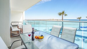 Ein Balkon mit Glasgeländer, ein Tisch mit einer Weinflasche, Obst und einem Glas, sowie Blick auf Strand und Meer.
