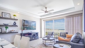 A spacious living area with a large window offering a view of the beach, a ceiling fan, and a dining area with a table and chairs. - Spectacular 2 Bedroom Condominium Encantame E102 (Puerto Peñasco)