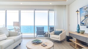 A modern living room with a sofa, armchairs, a coffee table, and a TV stand. Large windows offer a view of the ocean. - Encantame Towers Velero 2105 (Puerto Peñasco)