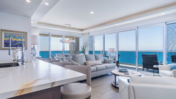 A modern living room with a large window overlooking the sea, a dining area with a white table, and a kitchen with a marble countertop. - Encantame Towers Velero 2105 (Puerto Peñasco)