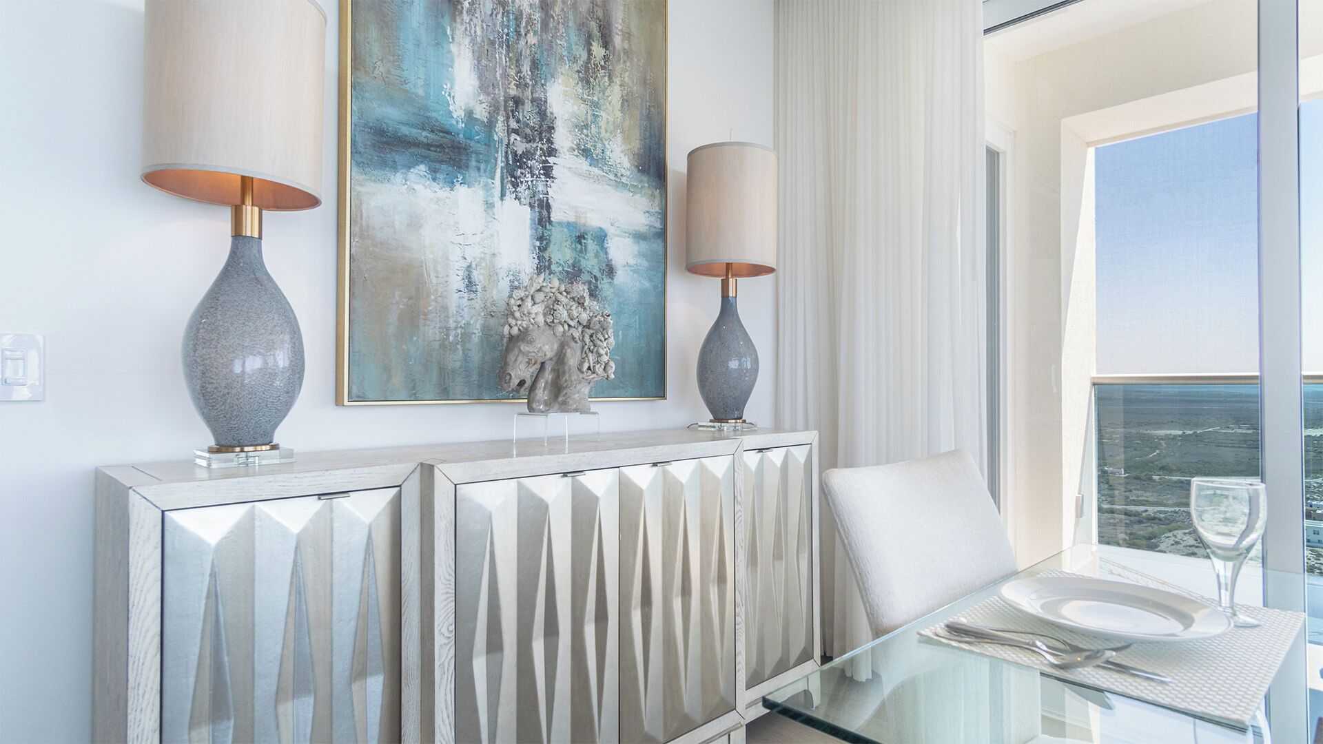 A modern dining area with a glass table, two wall-mounted lamps, a white console table, and a large abstract painting above.