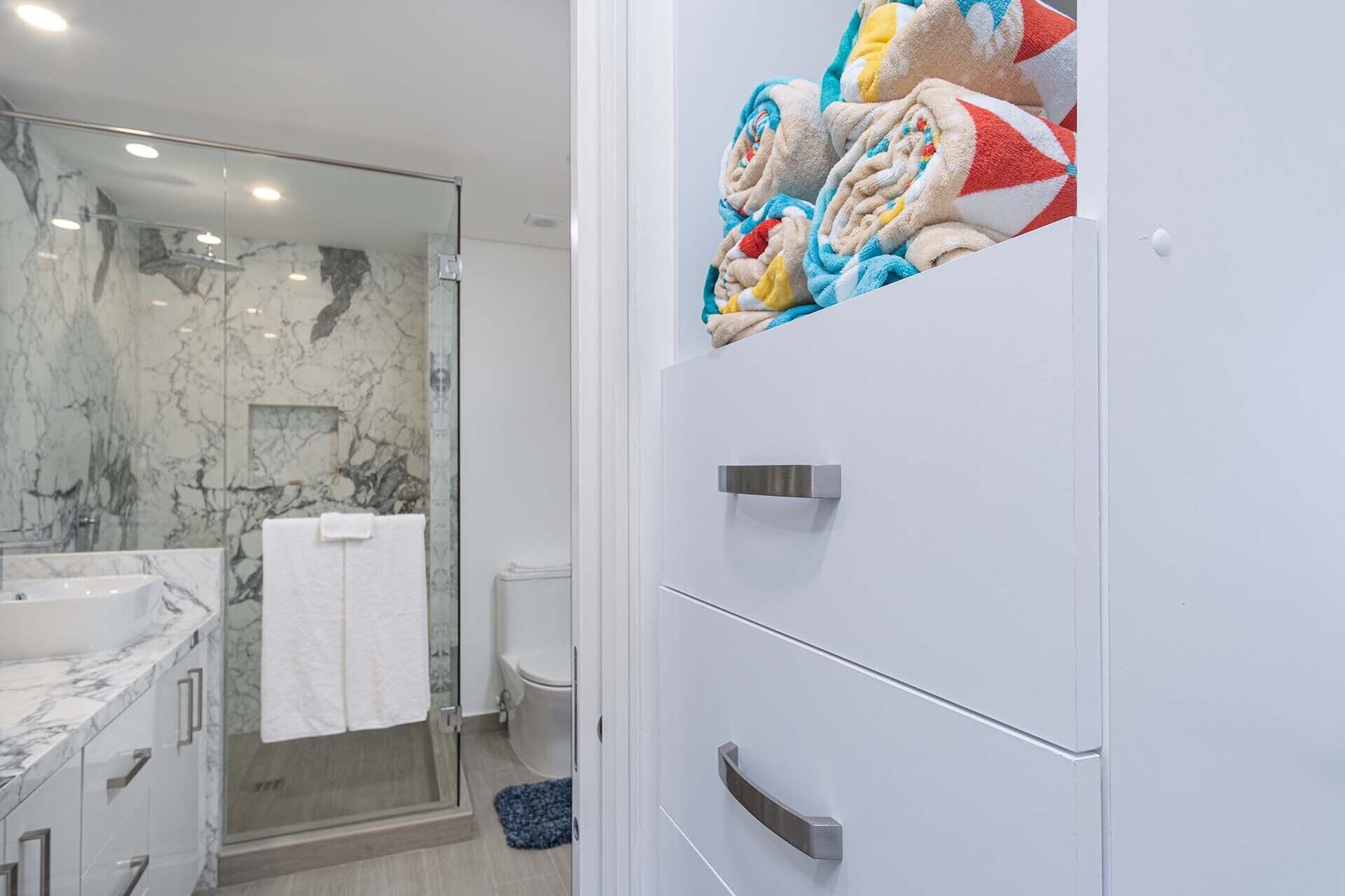 A bathroom with a glass shower enclosure, a marble countertop, and a towel rack with folded towels.