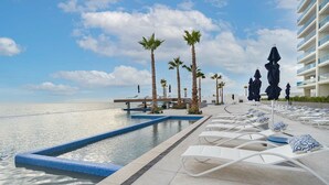 A poolside area with lounge chairs and umbrellas, palm trees, and a view of the sea. - Sublime 1 Bedroom Condo Encantame Towers Verano 503 (Puerto Peñasco)