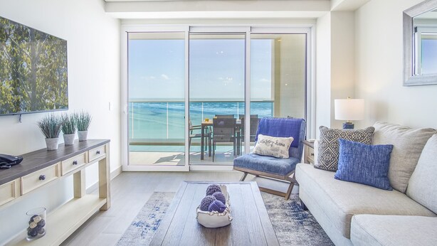 A modern living room with a large sliding glass door leading to a balcony with a view of the ocean. There is a sofa, a coffee table, and a TV on the wall. - Sublime 1 Bedroom Condo Encantame Towers Verano 503 (Puerto Peñasco)