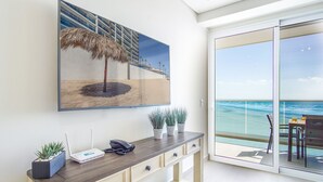 A modern room with a large beach-themed painting, a wooden console table with a phone and a projector, and a balcony with a view of the ocean. - Sublime 1 Bedroom Condo Encantame Towers Verano 503 (Puerto Peñasco)