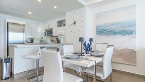 A modern kitchen with white cabinetry, a stainless steel refrigerator, and a microwave. There is a dining area with white chairs and a table set for two. A large painting of the ocean is on the wall. - Amazing Condo by the Beach Encantame W1203 (Puerto Peñasco)
