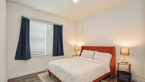 Ein Schlafzimmer mit einem Bett, zwei Nachttischen mit Lampen, einem Fenster mit Vorhängen und Holzdielen.