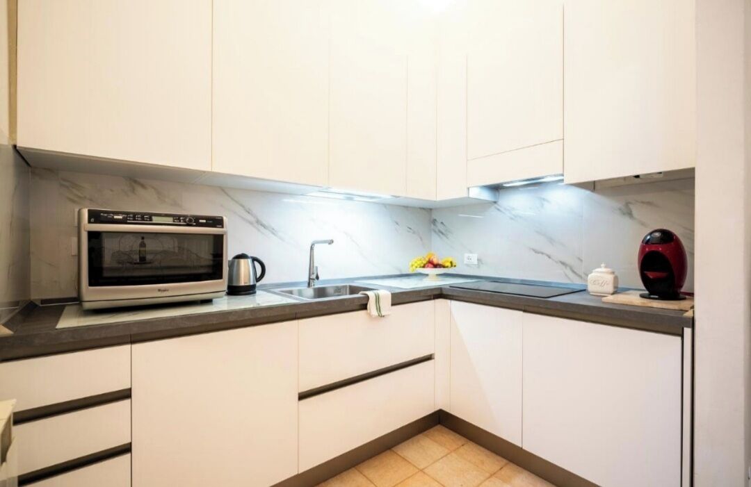 A compact kitchen with white cabinets, a built-in microwave, a kettle, and a sink.