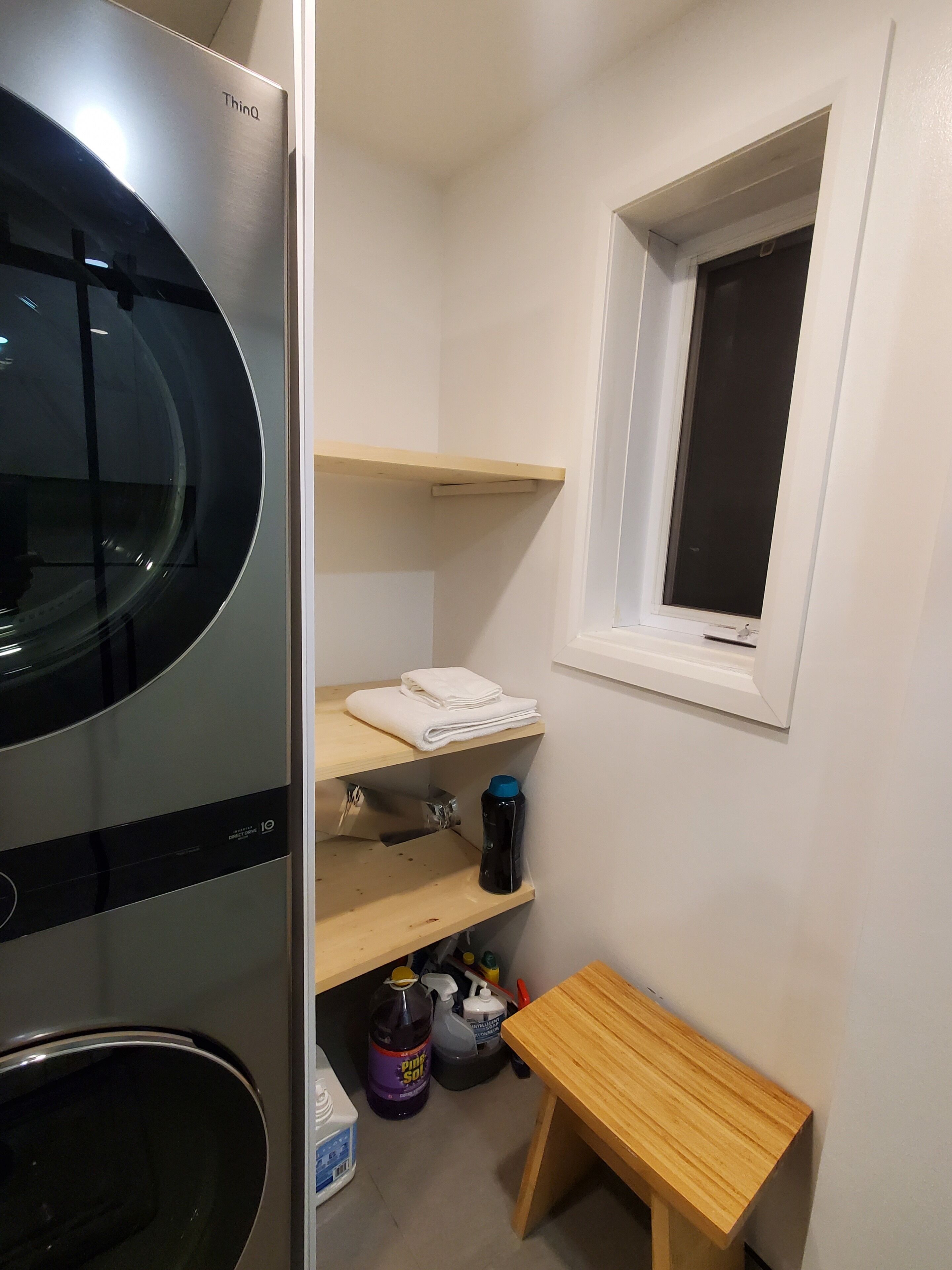 A compact kitchen with a built-in refrigerator, a microwave, and a sink. There are wooden shelves above the sink and a small wooden table beside it. A window with a black blind is visible on the wall.