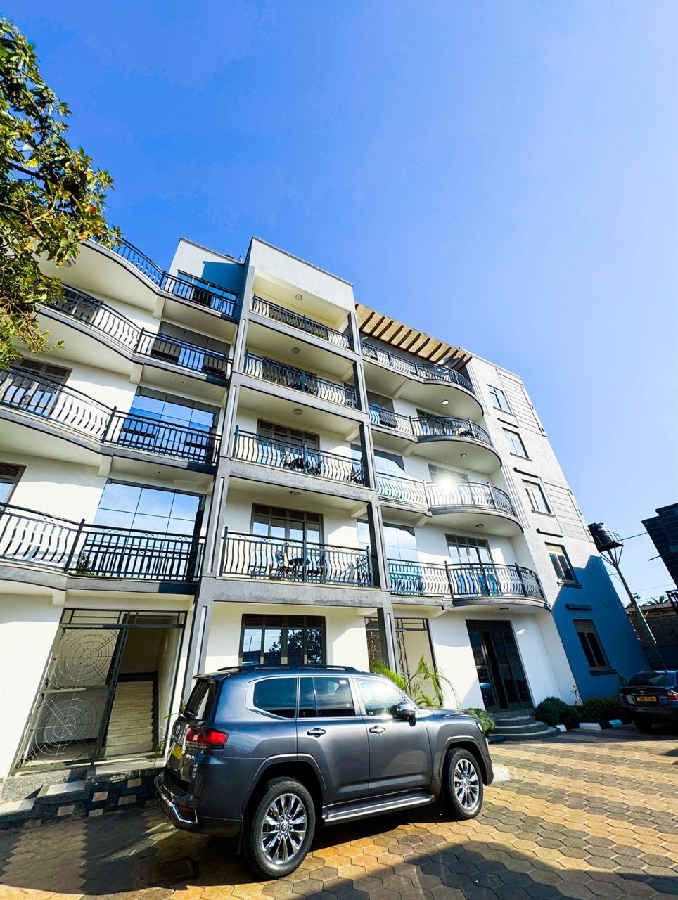A multi-story residential building with balconies, a parked SUV, and a clear blue sky.