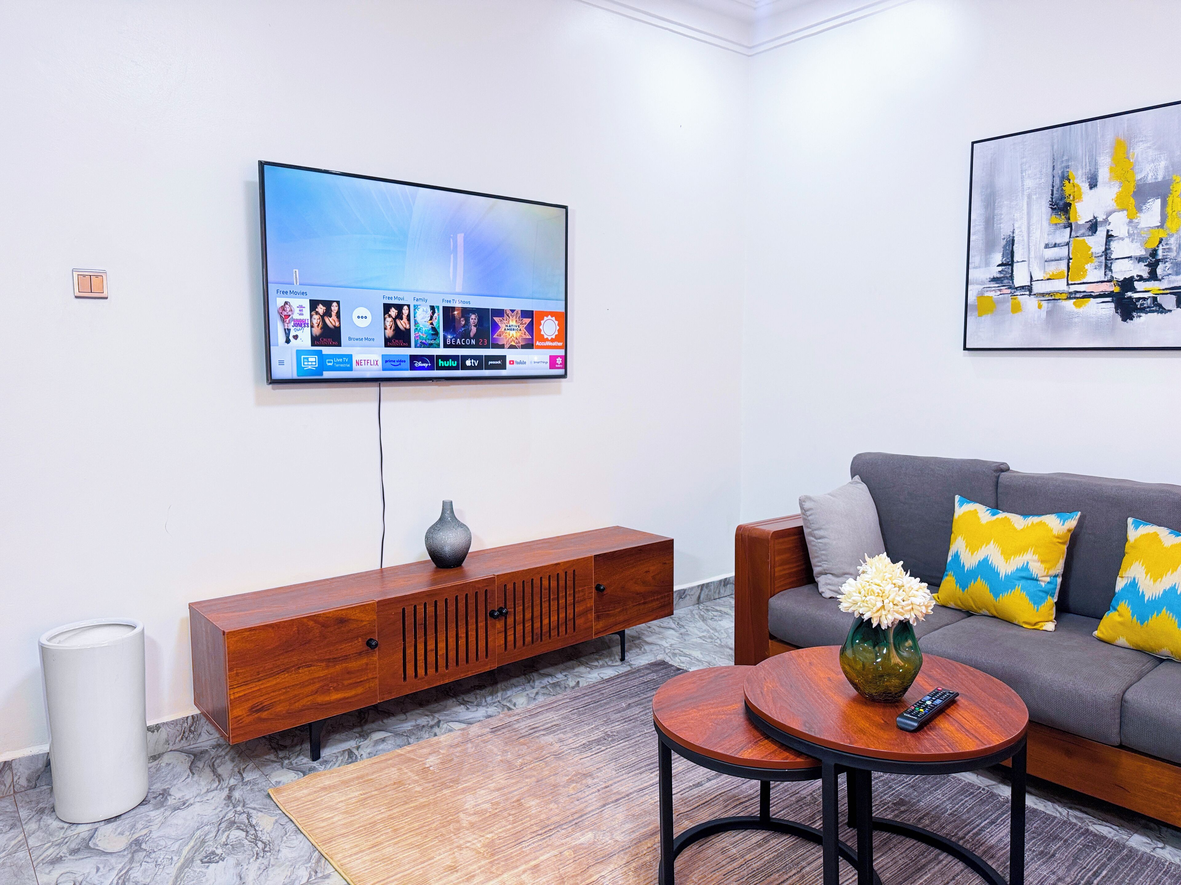A modern living room with a flat-screen TV mounted on the wall, a wooden console table, a grey sofa with patterned cushions, and a small round table with a vase and remote control.