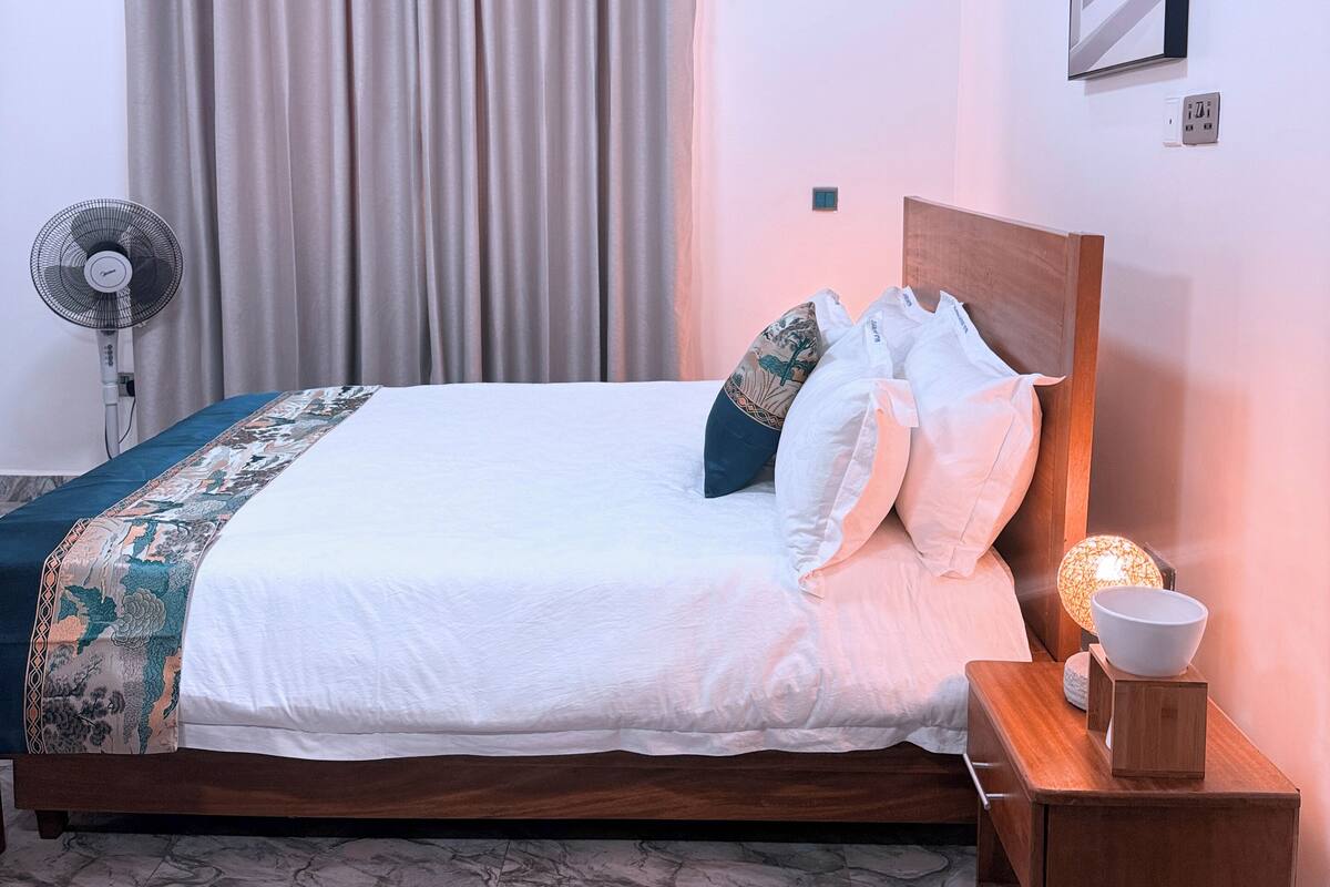 Un lit bien fait, avec une tête de lit en bois, une table de chevet sur laquelle se trouve un bol, et une lampe fixée au mur.