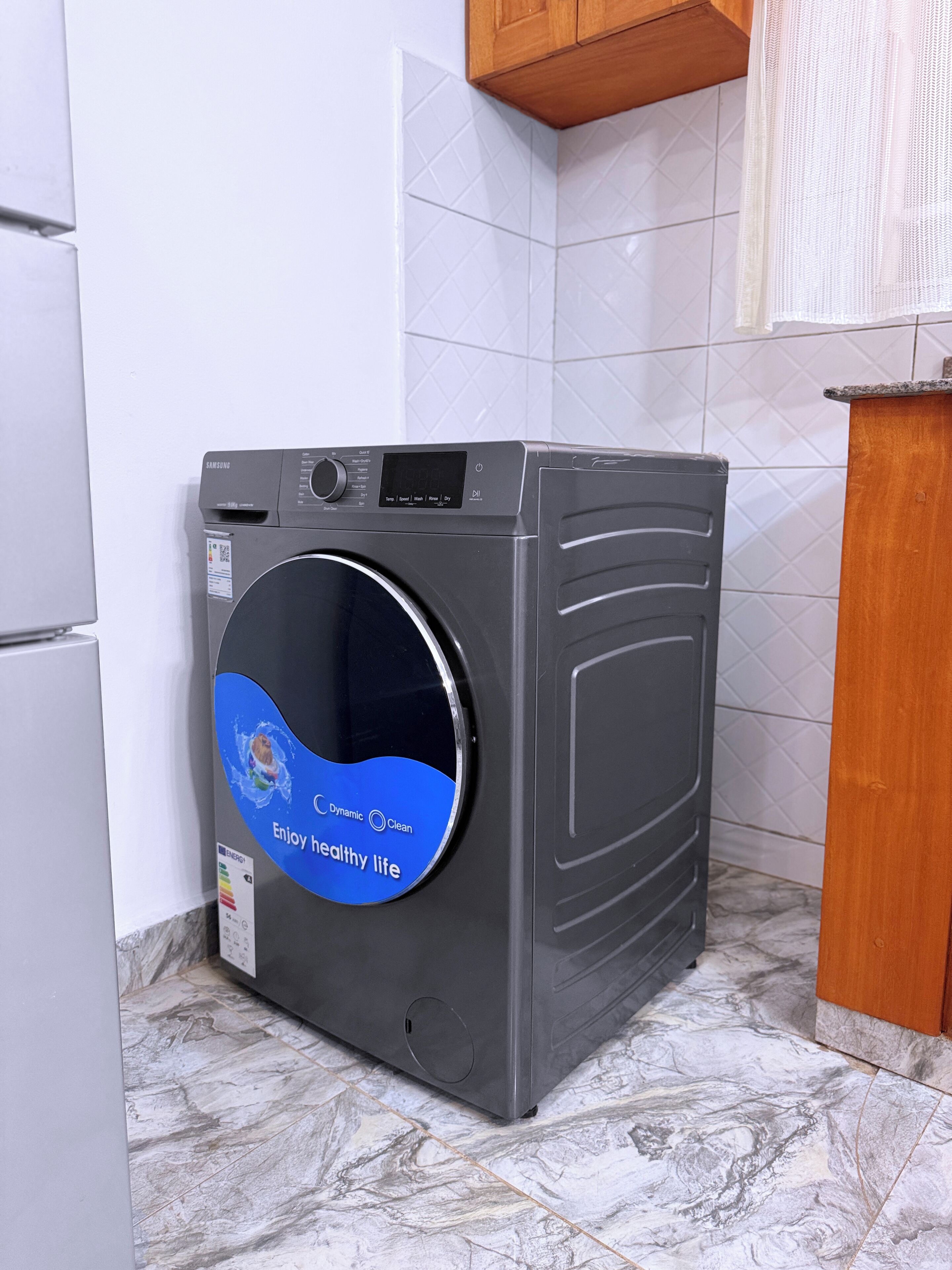 A front-loading washing machine in a kitchen with white tiled walls and a marble floor.