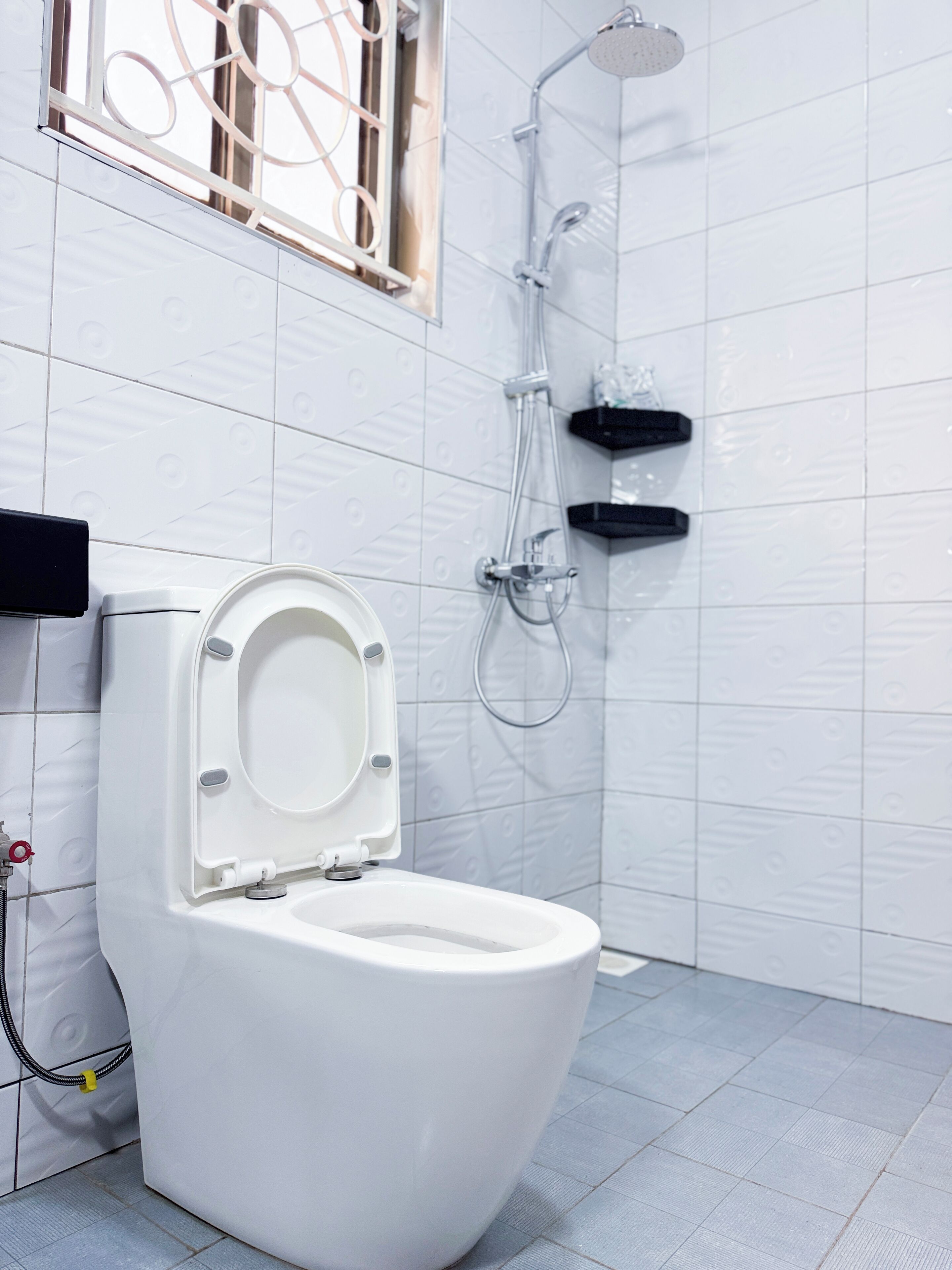 Un baño moderno con inodoro blanco, ducha con cabezal de mano y una ventana con celosías decorativas.
