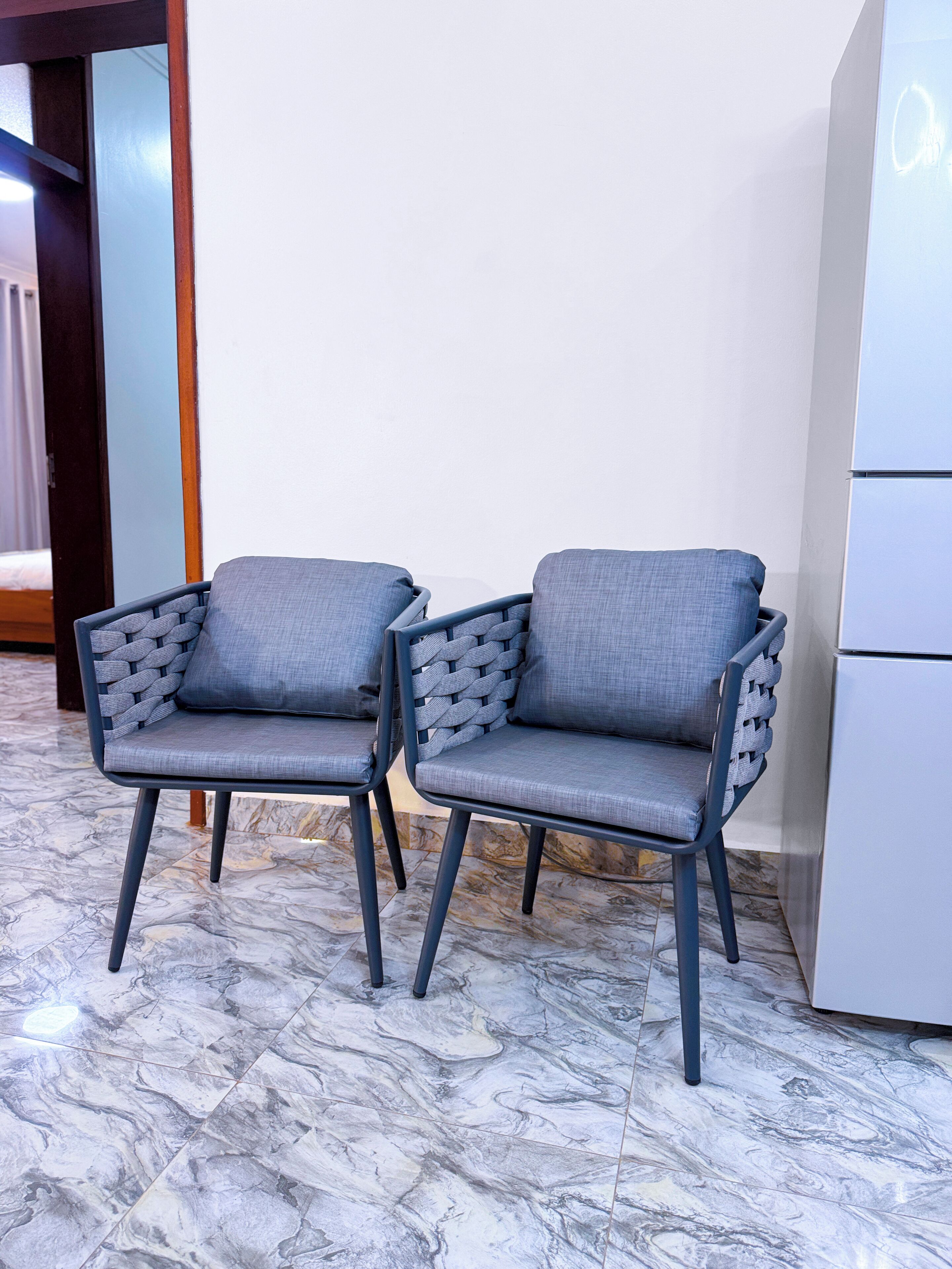 Deux fauteuils gris, recouverts de tissu, avec des accoudoirs en osier, sont placés sur un sol en marbre dans une pièce dont le mur est blanc et où l’on aperçoit, à l’arrière-plan, un appareil électroménager blanc.