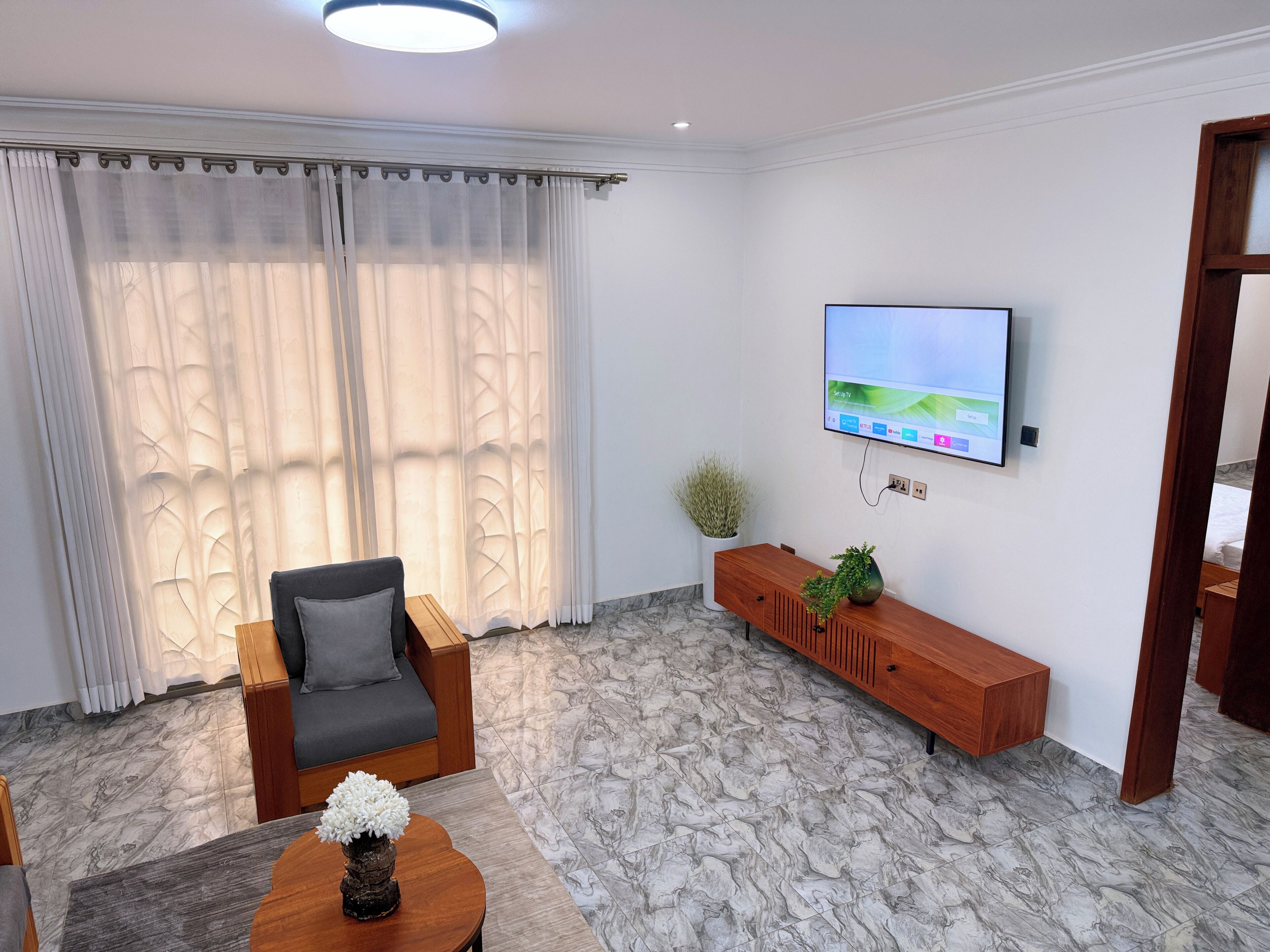 A modern living room with a grey sofa, a wooden coffee table with a white flower vase, a TV mounted on the wall, and a marble-patterned floor.