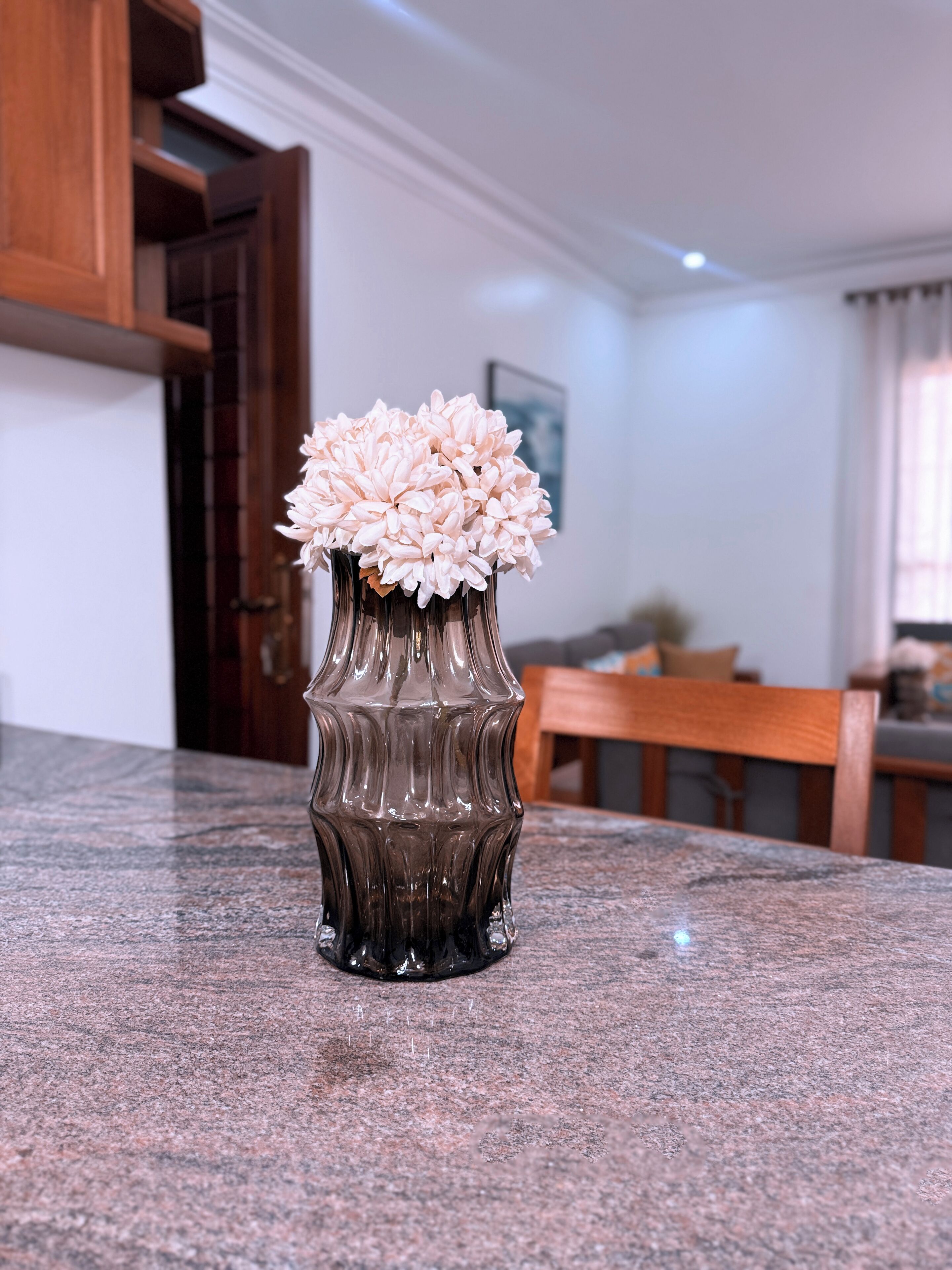 Eine Küche mit einer Granitabdeckung, einer dunklen Glasvase mit rosa Blumen, Holzschränken und einem Essbereich im Hintergrund.