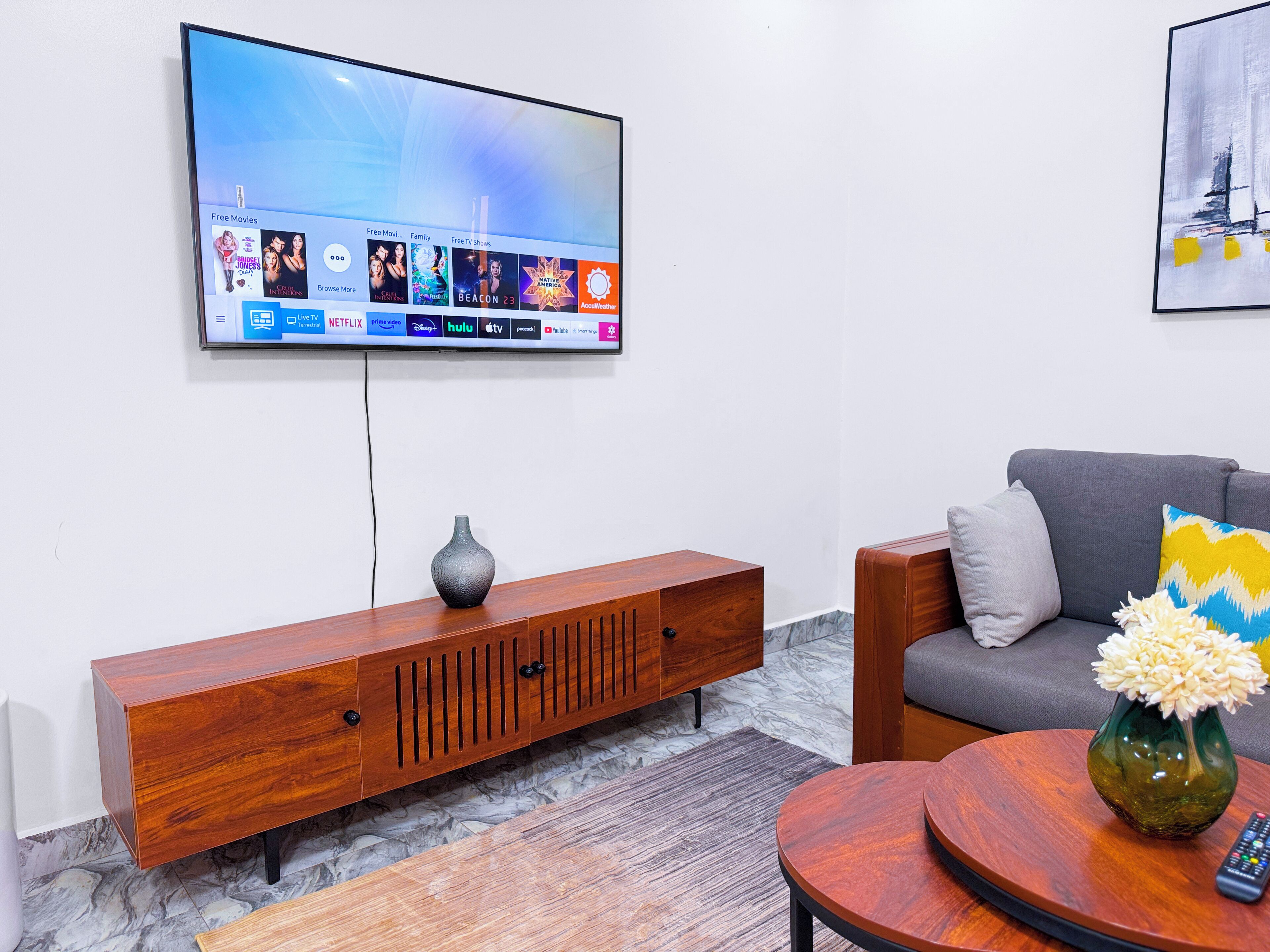 A living room with a flat-screen TV mounted on the wall, a wooden console table, a grey sofa, and a round wooden coffee table with a vase and remote control.