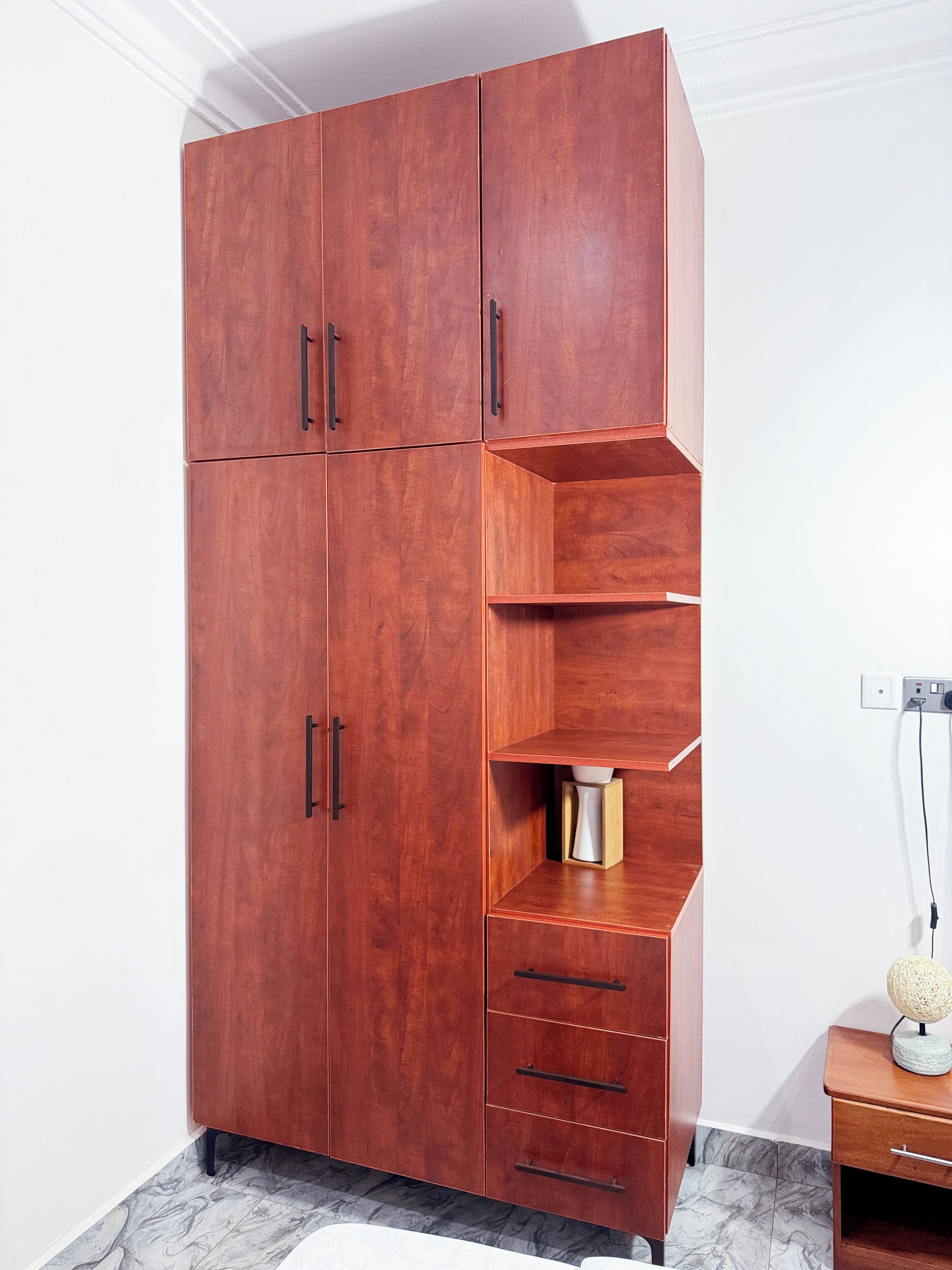 A wooden wardrobe with multiple compartments and drawers, a small shelf unit, and a power outlet on the wall.