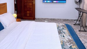 Ett sovrum med en säng, en tv som är monterad på väggen, en golvfläkt och en dörr av trä.