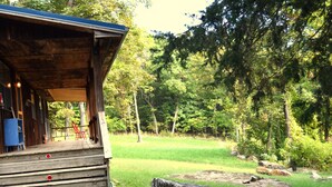 A wooden cabin with a porch, surrounded by a forested area with a grassy field and rocks.