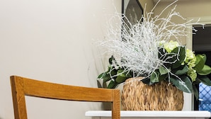A wooden chair with a cushioned seat and backrest, positioned in front of a white table and a woven basket containing green plants and white branches. - Caffarella Vacanze, comfort and relaxation a few minutes from the heart of Rome (Roma)