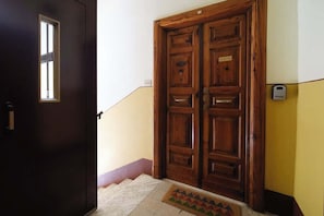 A wooden double door with a small window, a doormat, and a wall-mounted device.