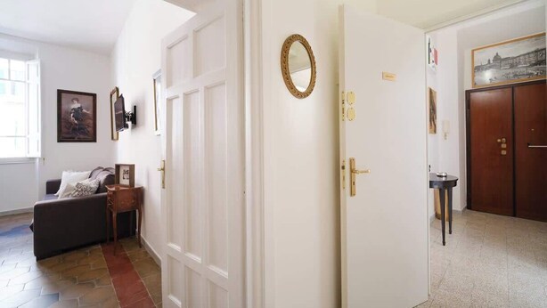 A hallway with a white door, a framed mirror, a small table with a lamp, and a couch. - BuddyRent - Vatican Domus - 4 ospiti (Rome)