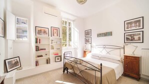 Ein Schlafzimmer mit einem Bett, Nachttisch, Lampe und an der Wand befestigten Regalen mit Büchern und Bildern.
