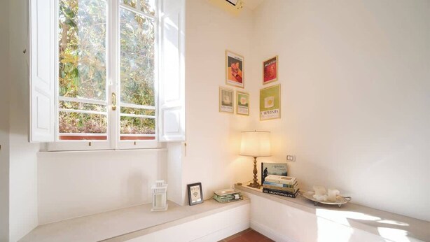 Ein Zimmer mit Fenster, einer Lampe, einem Regal mit Büchern und gerahmten Bildern an der Wand.